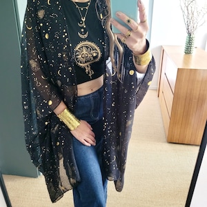 Może przedstawiać: Czarny, przezroczysty kimono ze złotym wzorem niebieskim jest noszony na czarnym topie z grafiką słońca i księżyca. Strój zawiera niebieskie dżinsy i złotą biżuterię, w tym bransoletkę i pierścionki. Ogólnym tematem jest boho.