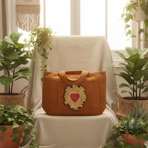 Könnte beinhalten: Eine Cord-Tragetasche in einem warmen Orangeton, mit einem herzförmigen Emblem mit goldenen und roten Akzenten. Die Tasche hat eine rechteckige Form mit einer Tasche und Griffen. Das Bild ist in einer hellen, natürlichen Umgebung mit Pflanzen aufgenommen.
