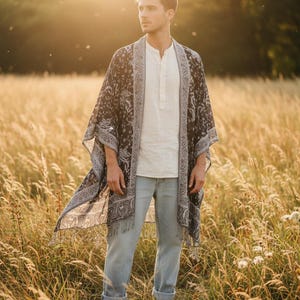 Könnte beinhalten: Ein dunkler Kimono mit Paisley-Muster und Fransenbesatz wird über einem weißen Hemd und hellblauen Jeans getragen. Die Person steht in einem Feld mit hohem Gras, mit der untergehenden Sonne im Hintergrund.