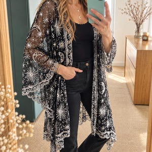 Peut inclure: Un kimono noir transparent orné de broderies complexes en sequins argentés représentant des motifs célestes, notamment des étoiles et des croissants de lune. Le vêtement présente des manches larges et fluides ainsi qu'un ourlet festonné.