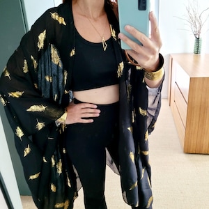 Könnte beinhalten: Schwarzer Kimono mit goldenem Federdruck. Der transparente Stoff wird über einem schwarzen Top und Leggings getragen. Eine goldene Anhängerkette und ein Armband ergänzen den Look. Der Kimono ist eine stilvolle Ergänzung für jede Garderobe.