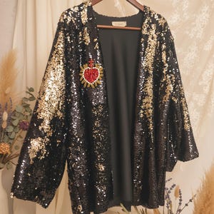 May include: Black sequin jacket with gold accents and a red heart emblem. The open-front jacket is hanging on a wooden hanger. The jacket is a statement piece, perfect for special occasions.