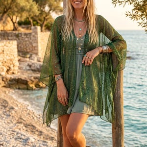 Può includere: Un kimono verde bosco trasparente con un sottile riflesso metallico e un delicato motivo a pois, indossato sopra un miniabito verde salvia a tinta unita. L'outfit è completato da collane dorate a strati, bracciali in metallo impilati e anelli.