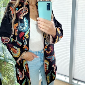 Könnte beinhalten: Eine lange schwarze Jacke mit bunter Blumenstickerei. Die Jacke hat einen Stehkragen und einen Reißverschluss vorne. Das Model trägt die Jacke zu einer blauen Jeans.