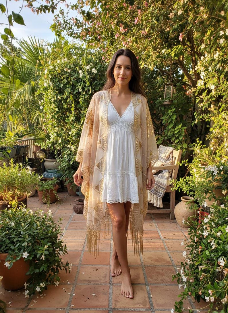 May include: A white sleeveless mini dress with a V-neckline and lace trim details, worn under a sheer, beige, open-front kimono featuring gold embroidered sunburst patterns and long fringe along the bottom hemline.