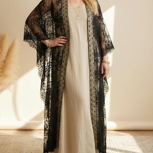 Op de afbeelding: Een zwarte kanten kimono-achtige cover-up met gouden rand over een beige jurk. De kimono heeft ingewikkelde bloemenkantpatronen en een vloeiend ontwerp. De jurk is een eenvoudige, lange, neutrale jurk. De outfit is geaccessoireerd met gouden sieraden.
