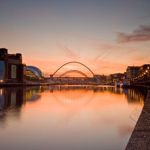 Puede incluir: Una vista de un puente moderno sobre un río al atardecer. El puente es una estructura de arco de acero con un diseño curvo. El río está tranquilo y refleja los tonos naranja y rosa del cielo del atardecer. Los edificios bordean la orilla del río a ambos lados.