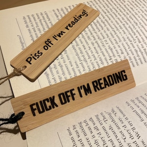 May include: Two bamboo bookmarks with black text. One says "Piss off I'm reading!" and the other says "Fuck off I'm reading". Both have a hole with twine for attaching to a book. The bookmarks are on top of an open book.