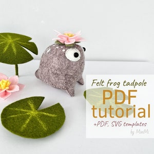 May include: Felt frog tadpole PDF tutorial with PDF and SVG templates by MiniMi. The image shows a gray felt frog with a pink flower on its head, sitting on a white surface with green felt lily pads.