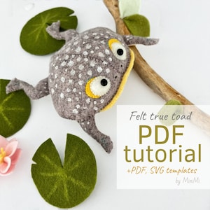 Könnte beinhalten: Ein grauer Filz-Kröte mit weißen Punkten und gelben Augen sitzt auf einem Ast mit grünen Filz-Seerosen. Der Text "Felt true toad PDF tutorial +PDF, SVG templates by MiniMi" befindet sich unter der Kröte.