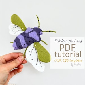 Peut inclure: Un insecte puant en feutre violet et vert avec des ailes blanches. L'insecte est tenu dans une main. Le texte "Felt lilac stink bug PDF tutorial +PDF, SVG templates by MiniMi" est en arrière-plan.