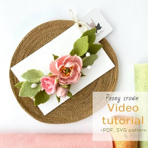 May include: A pink felt peony flower crown with green leaves and white felt accents. The crown is displayed on a brown woven mat with a white card that says "Peony crown Video tutorial +PDF, SVG pattern by MiniMi".