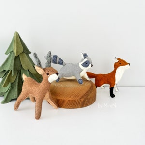 May include: A set of four felt woodland animal toys: a brown deer, a grey raccoon, and an orange fox. The toys are arranged on a wooden platform in front of a green felt Christmas tree. The text "by MiniMi" is visible in the bottom right corner of the image.