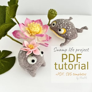 May include: A PDF tutorial for a swamp life project featuring two gray felt frogs with white eyes and yellow accents, one with a pink flower on its head, and a pink lotus flower. The text "Swamp life project PDF tutorial +PDF, SVG templates by MiniMi" is displayed on a white background.