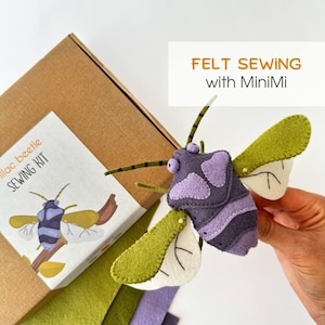 May include: A felt sewing kit with a brown cardboard box featuring the text 'Lilac beetle SEWING KIT' and 'FELT SEWING with MiniMi'. A handcrafted purple and green felt beetle toy with stitched details and antennae is held in the foreground.