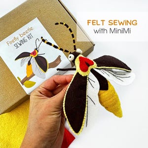 May include: A felt sewing kit for a firefly beetle, featuring a brown cardboard box with a label reading Firefly beetle SEWING KIT and FELT SEWING with MiniMi. The kit includes a finished felt firefly beetle with dark brown wings, white underwings, a yellow abdomen, and black and yellow antennae.