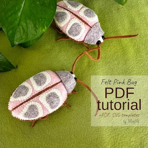 May include: Two handmade felt bugs in pink, gray, and brown, with long antennae, are displayed on a green surface. The bugs have white and gray patterned wings. The image includes text that reads "Felt Pink Bug PDF tutorial + PDF, SVG templates by MiniMi."