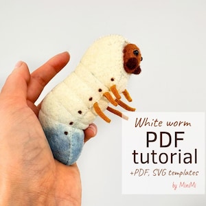 Puede incluir: Un gusano blanco de fieltro con patas marrones y naranjas y una cola azul. El gusano está siendo sostenido en una mano. El texto "White worm PDF tutorial +PDF, SVG templates by MiniMi" está en la esquina inferior derecha de la imagen.