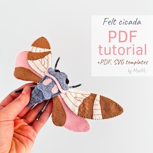 Può includere: Tutorial di artigianato di cicala in feltro con modelli PDF e SVG. L'immagine mostra una mano che tiene una cicala in feltro finita con feltro marrone, rosa, bianco e grigio.