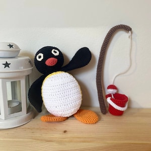 Puede incluir: Un juguete de pingüino de crochet con colores negro y blanco, patas naranjas y un pico amarillo. El pingüino sostiene una caña de pescar marrón con un cubo rojo al final.