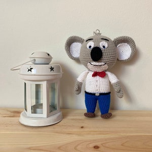 Puede incluir: Un juguete de peluche de koala tejido a crochet que lleva una camisa blanca con una corbata de lazo roja y pantalones azules. El juguete está de pie sobre una superficie de madera junto a una linterna blanca.