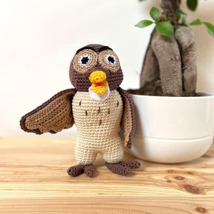 Peut inclure: Un jouet hibou en crochet fait de fil brun et beige, avec de grands yeux noirs, un bec jaune et des accents blancs. Le hibou est debout sur une surface en bois à côté d'une plante en pot.