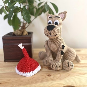 以下が含まれることがあります： 木製の表面に座っている、黒点のある茶色とベージュの毛糸で編まれた犬。犬は白い目と黒い鼻をしています。犬の左側の表面には、赤と白の毛糸で編まれたサンタの帽子があります。