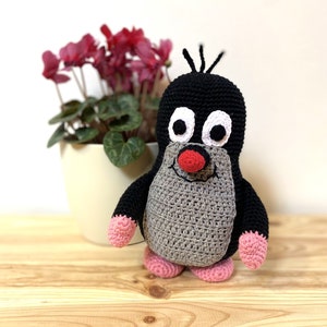 May include: A crocheted mole plush toy with black fur, grey body, pink feet, and a red nose. The toy has large white eyes and a friendly expression.