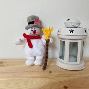 Puede incluir: Un juguete de mu&ntilde;eco de nieve de crochet que lleva un sombrero de copa gris con una flor rosa, una bufanda roja y sostiene una escoba marr&oacute;n y amarilla.