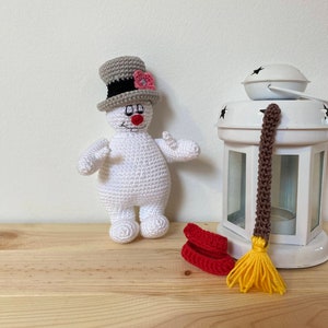 Puede incluir: Un mu&ntilde;eco de nieve de crochet blanco con un sombrero de copa gris y una flor rosa. El mu&ntilde;eco de nieve sostiene una escoba roja con cerdas amarillas.
