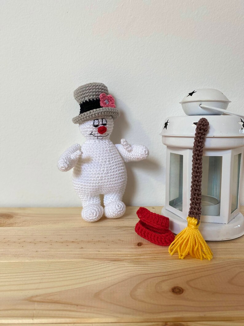 Puede incluir: Un mu&ntilde;eco de nieve de crochet blanco con un sombrero de copa gris y una flor rosa. El mu&ntilde;eco de nieve sostiene una escoba roja con cerdas amarillas.