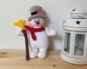 May include: A crocheted snowman toy with a gray top hat, red scarf, and a brown stick broom. The snowman is standing on a wooden surface.
