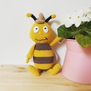 May include: A crocheted bee plush toy with a brown and yellow striped body, black eyes, and a white smile. The bee is wearing a brown hat and is sitting on a wooden surface.