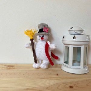 Puede incluir: Un mu&ntilde;eco de nieve de crochet blanco con un sombrero de copa gris, una flor rosa y una bufanda roja. El mu&ntilde;eco de nieve sostiene una escoba marr&oacute;n y amarilla.
