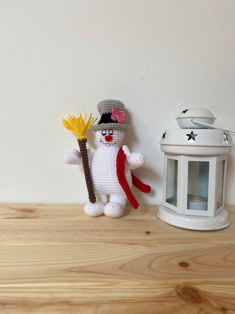 Puede incluir: Un mu&ntilde;eco de nieve de crochet blanco con un sombrero de copa gris, una flor rosa y una bufanda roja. El mu&ntilde;eco de nieve sostiene una escoba marr&oacute;n y amarilla.