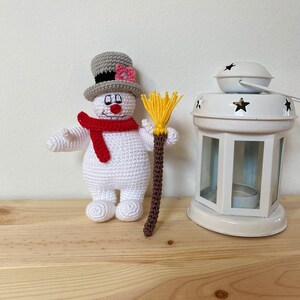Puede incluir: Un juguete de mu&ntilde;eco de nieve de crochet que lleva un sombrero de copa gris con una flor rosa, una bufanda roja y sostiene una escoba marr&oacute;n y amarilla.