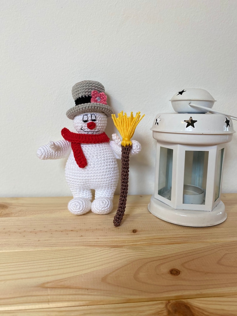 Puede incluir: Un juguete de mu&ntilde;eco de nieve de crochet que lleva un sombrero de copa gris con una flor rosa, una bufanda roja y sostiene una escoba marr&oacute;n y amarilla.