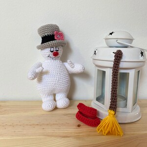 Puede incluir: Un mu&ntilde;eco de nieve blanco de crochet con un sombrero de copa gris y una flor rosa. El mu&ntilde;eco de nieve est&aacute; de pie sobre una superficie de madera junto a una linterna blanca con una escoba marr&oacute;n y amarilla.