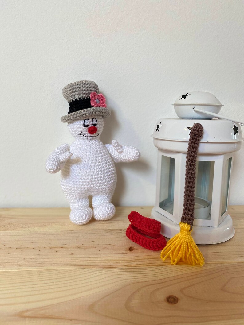 Puede incluir: Un mu&ntilde;eco de nieve blanco de crochet con un sombrero de copa gris y una flor rosa. El mu&ntilde;eco de nieve est&aacute; de pie sobre una superficie de madera junto a una linterna blanca con una escoba marr&oacute;n y amarilla.