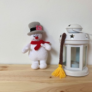 Puede incluir: Un mu&ntilde;eco de nieve de crochet con un sombrero gris, una bufanda roja y una flor rosa. El mu&ntilde;eco de nieve est&aacute; de pie sobre una superficie de madera junto a una linterna blanca con un borla marr&oacute;n y amarilla.
