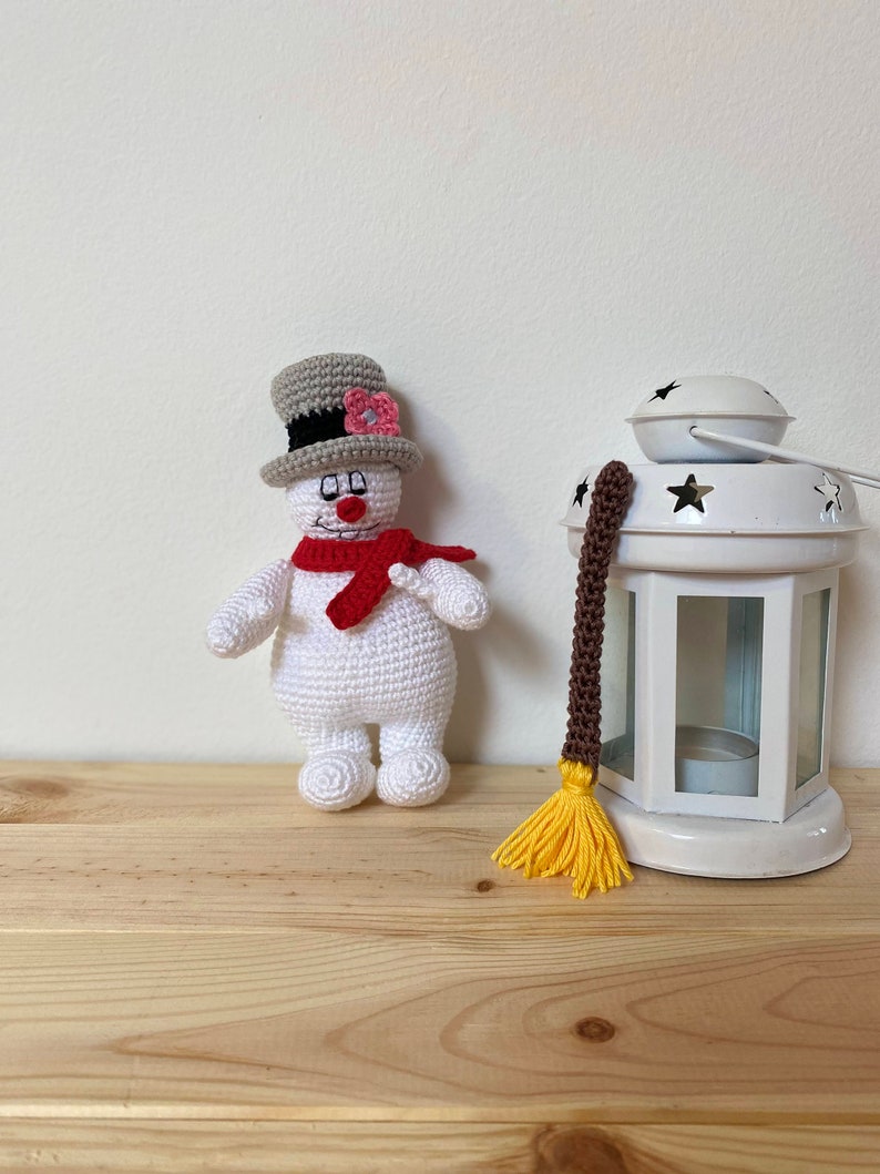 Puede incluir: Un mu&ntilde;eco de nieve de crochet con un sombrero gris, una bufanda roja y una flor rosa. El mu&ntilde;eco de nieve est&aacute; de pie sobre una superficie de madera junto a una linterna blanca con un borla marr&oacute;n y amarilla.
