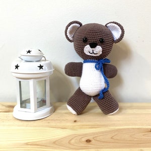 Op de afbeelding: Een bruine gehaakte teddybeer met witte accenten en een blauw lint om zijn nek. De beer staat op een houten oppervlak naast een witte lantaarn met zwarte sterren.