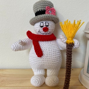 Puede incluir: Un mu&ntilde;eco de nieve de crochet con un sombrero de copa gris, una bufanda roja y una escoba marr&oacute;n. El mu&ntilde;eco de nieve tiene una flor rosa en su sombrero y una cara amigable.