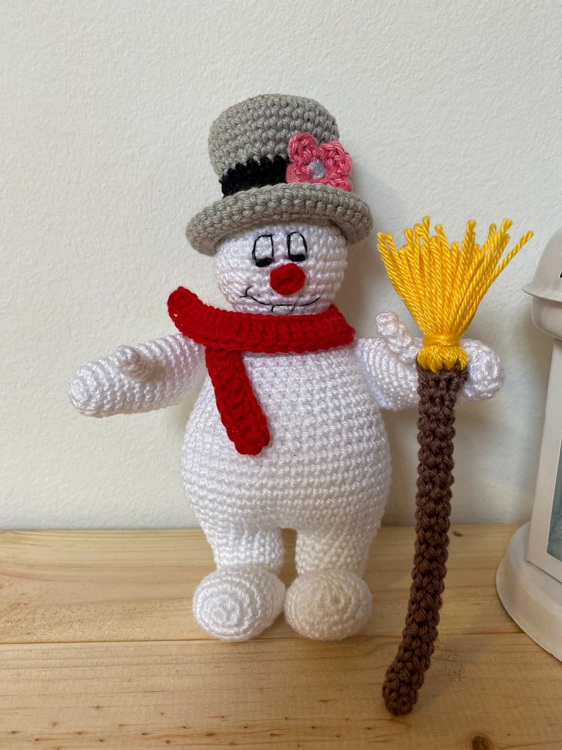 Puede incluir: Un mu&ntilde;eco de nieve de crochet con un sombrero de copa gris, una bufanda roja y una escoba marr&oacute;n. El mu&ntilde;eco de nieve tiene una flor rosa en su sombrero y una cara amigable.