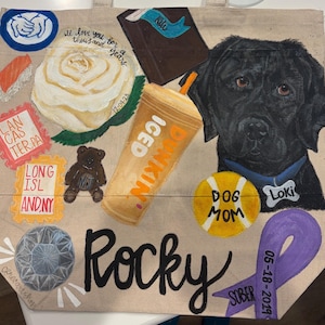 Puede incluir: Un bolso tote beige con un retrato de labrador negro y el nombre "Rocky". La bolsa presenta varios elementos pintados, incluyendo una rosa, sushi, un diamante y el texto "Dog Mom".