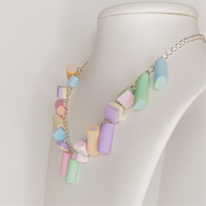 Pode incluir: Um colar de cor pastel com uma corrente prateada e vários pingentes em forma de doces. Os pingentes são em tons de rosa, azul, verde e amarelo.