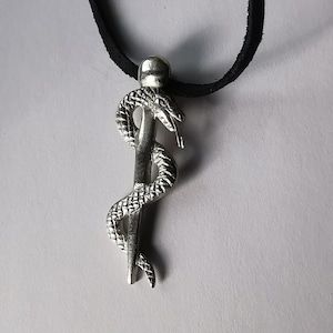 Puede incluir: Un collar con un colgante de plata que representa una serpiente enroscada alrededor de un bastón. El collar está en un cordón negro.
