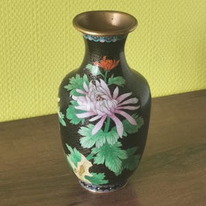 May include: Black decorative vase with a floral design. The vase features a gold rim and a black body adorned with a large pink and white flower, green leaves, and an orange flower. The vase has a textured surface.