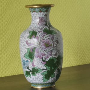 May include: A decorative vase with a floral design. The vase has a white background with a repeating pattern, and is adorned with a large pink and purple flower, green leaves, and blue accents. The rim is gold-coloured.