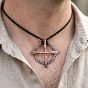 May include: A hammered copper pendant in the shape of a cross centered within a circle, suspended from a dark brown leather cord. The metal surface features a textured, rustic finish with visible indentations across the entire piece.